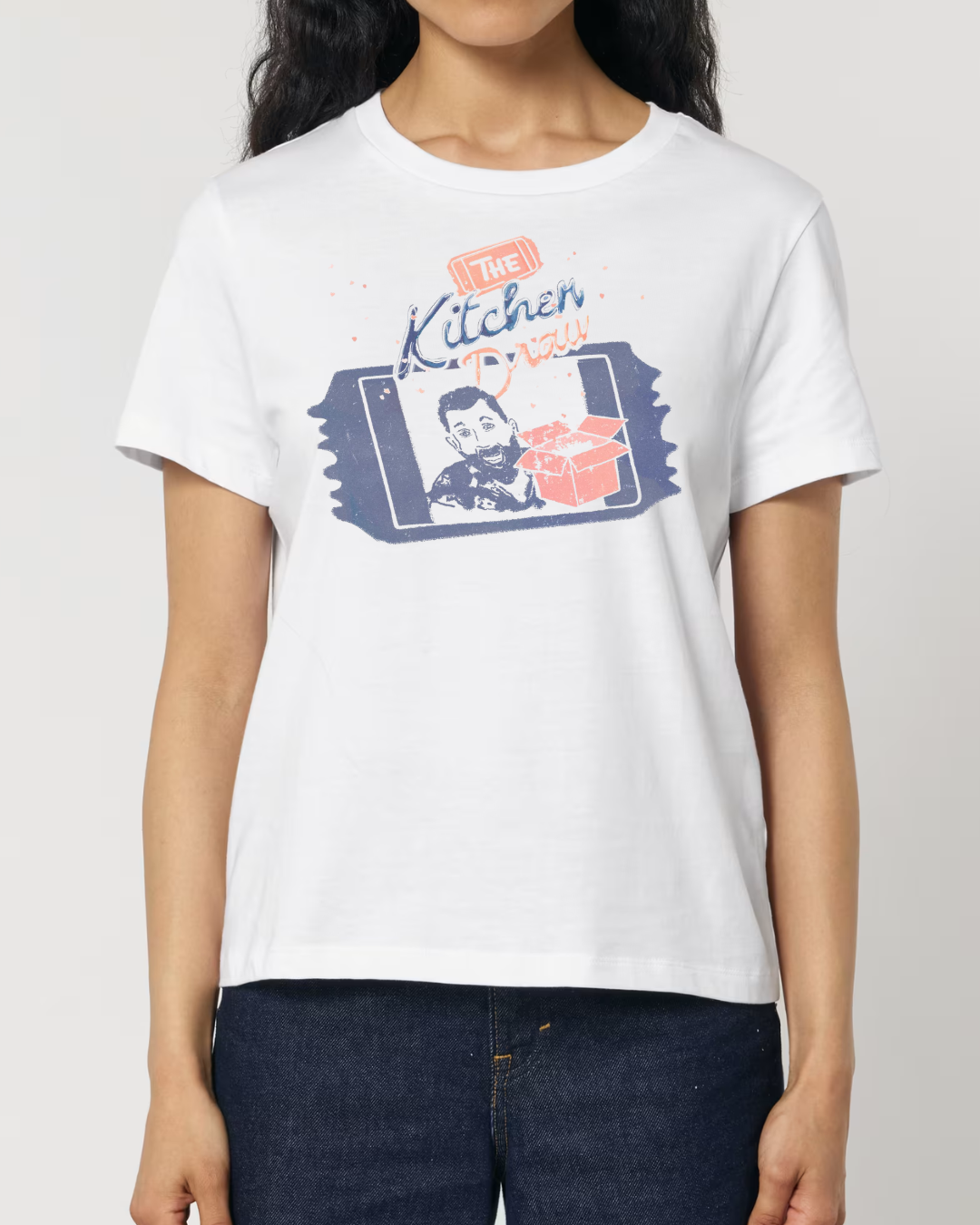 A woman wearing a white short-sleeve t-shirt with a colourful graphic print on the front that reads 'The Kitchen Draw' and features a stylised portrait, set against a plain light grey background.